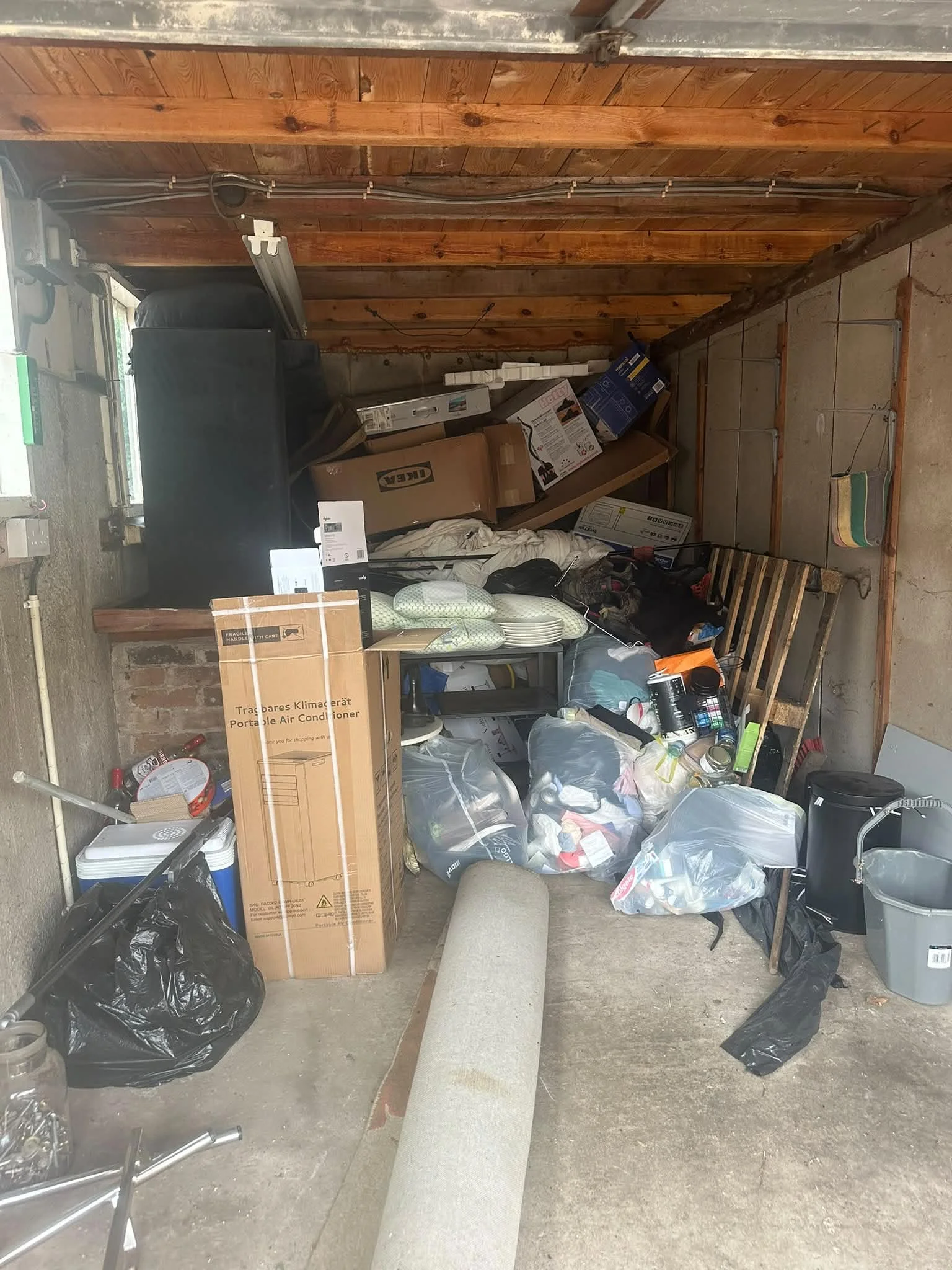 Single garage in York before clearance, packed with boxes, bagged rubbish, a rolled carpet, pallets and an old air conditioner