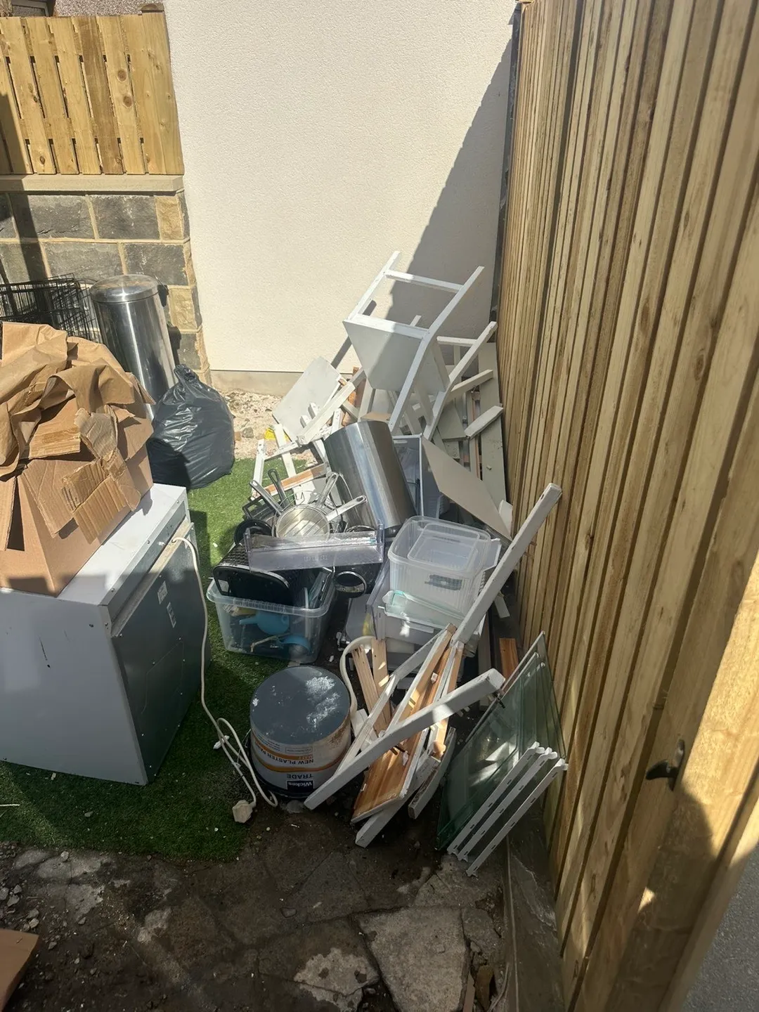 The same collection: the fridge at the edge of a pile of chairs, storage tubs, a paint tin and cardboard against a garden fence